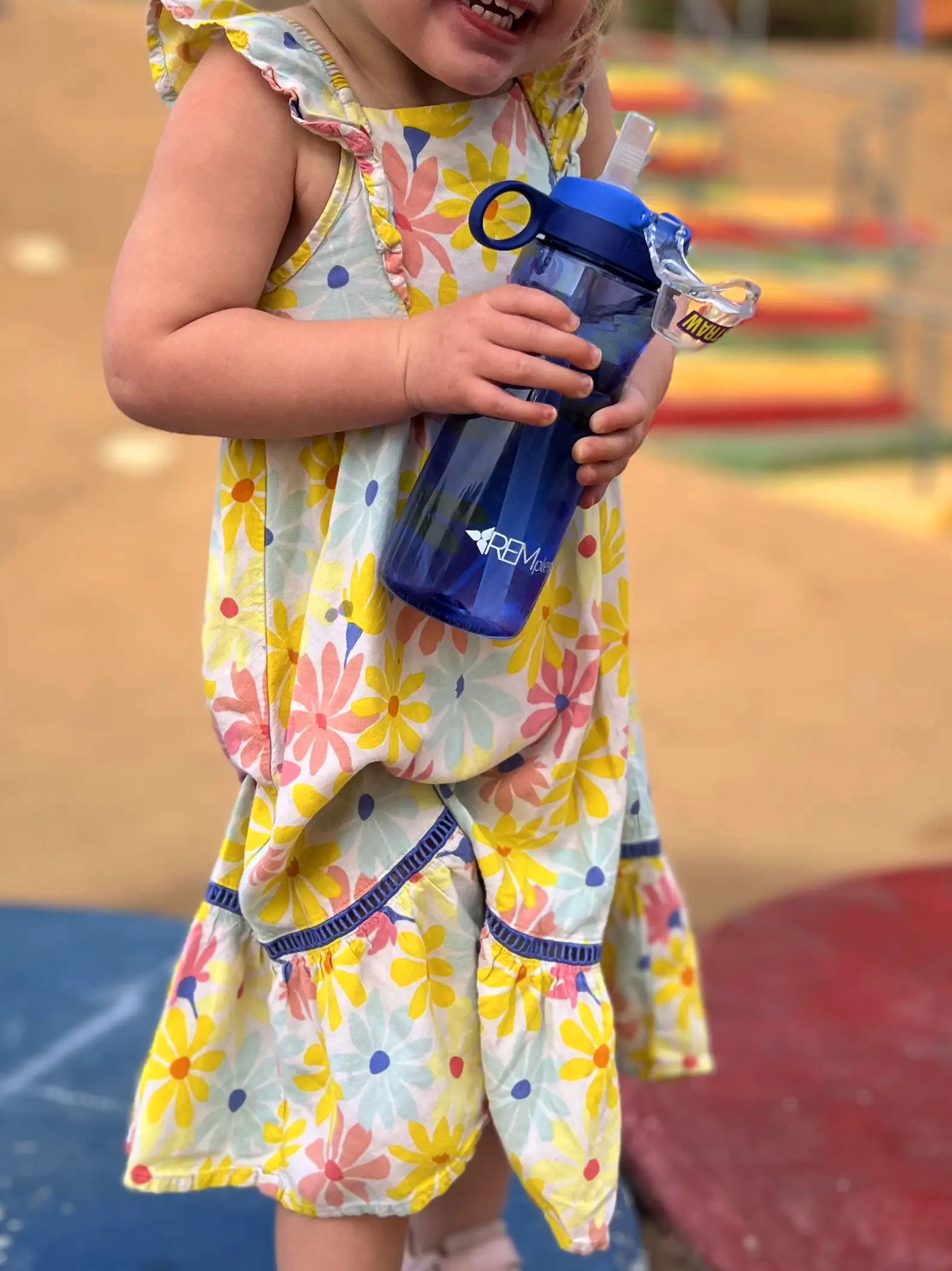 Child in a colorful dress holding a blue REMplenish water bottle on a playground.